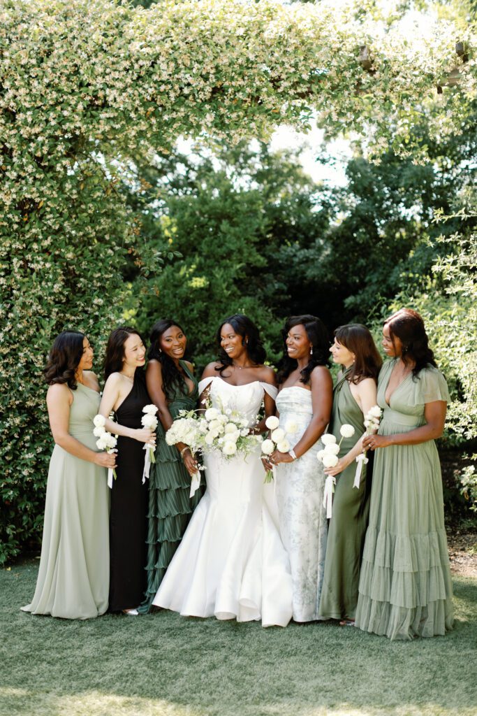 Bride in an off shoulder wedding gown standing with bridesmaids in emerald dresses holding white dahlia bouquets