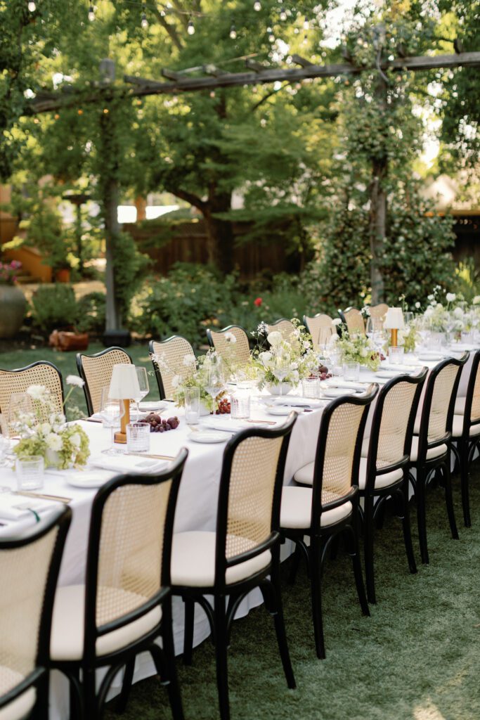 Long outdoor wedding reception table with string lights, black cane chairs, and white floral centerpieces at Tre Posti